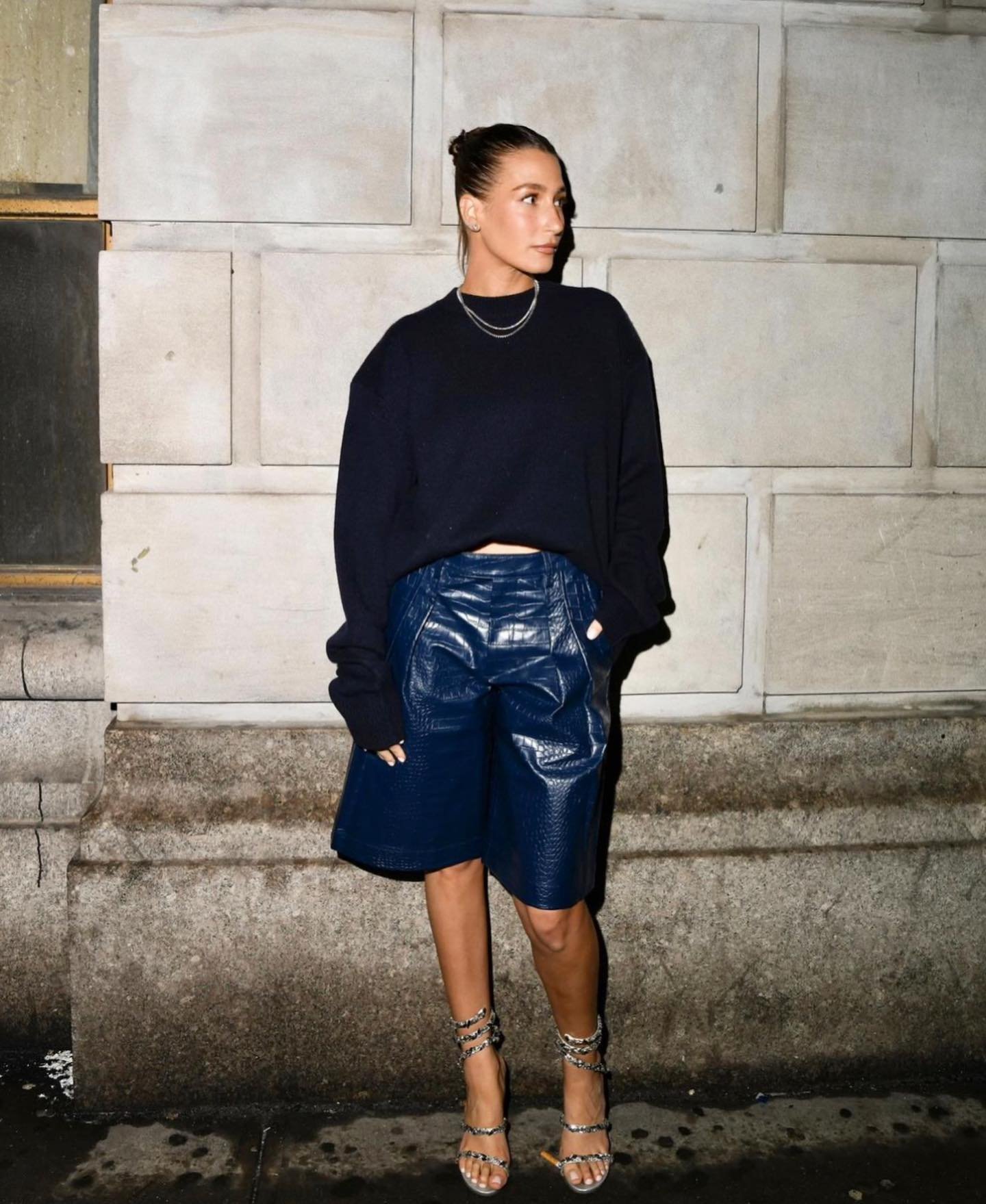 Fashionable adult woman poses against a stone wall wearing a black sweater, blue shorts, strappy heels, and jewelry.