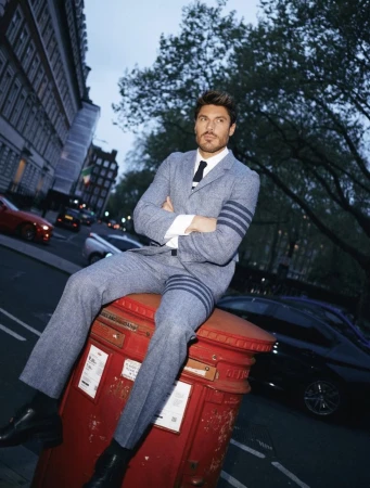 Man in a grey suit sitting on a red British post box in an urban environment, with apartment buildings and cars in the background.