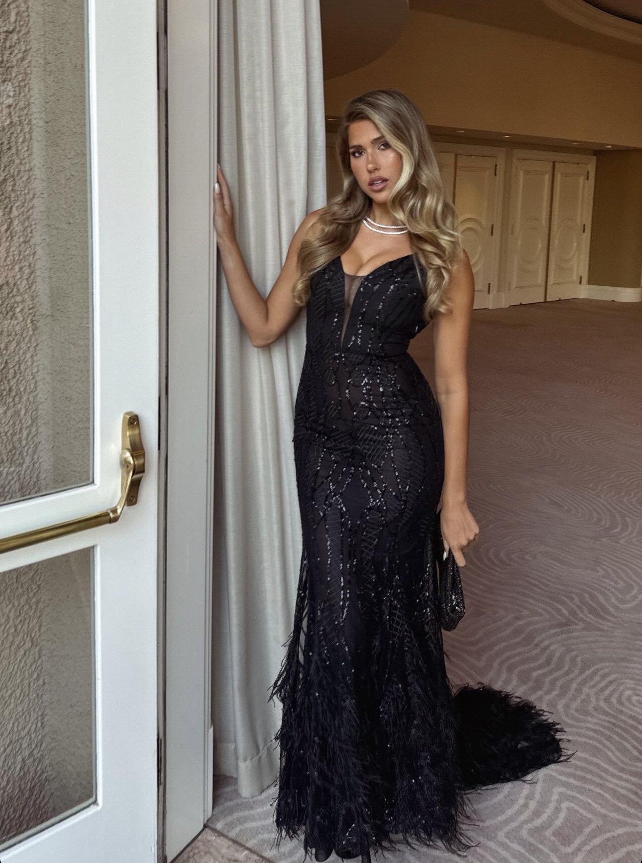 Woman in elegant black gown posing by door with neutral wall and carpet. Glamorous evening look, indoor setting, blonde hair.