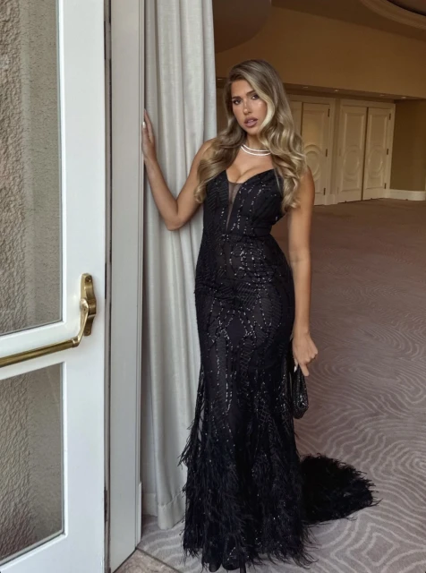 Woman in elegant black gown posing by door with neutral wall and carpet. Glamorous evening look, indoor setting, blonde hair.