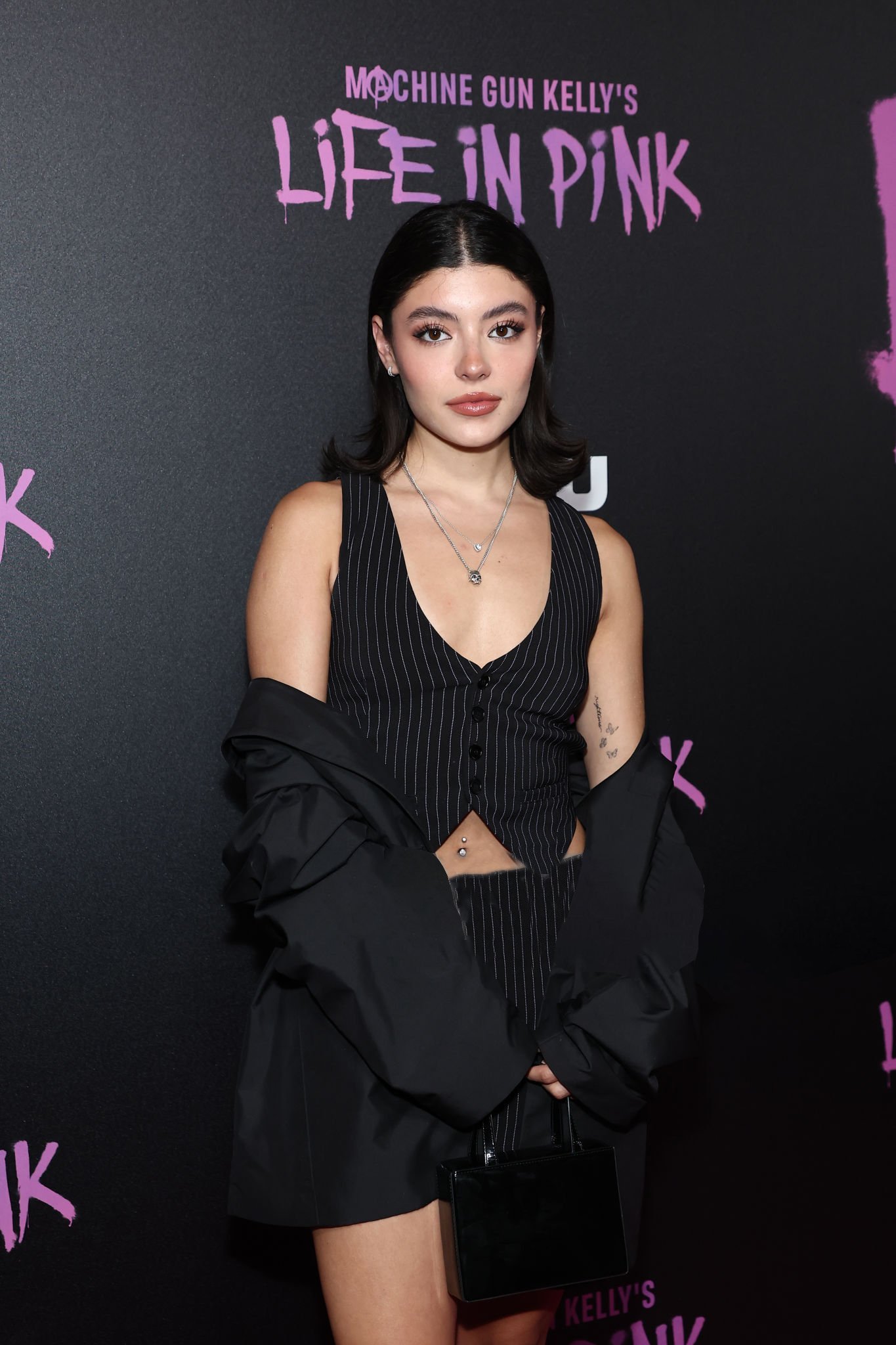 A fashionable young woman in a pinstripe vest and black jacket poses in front of a promotional backdrop. She holds a small purse.