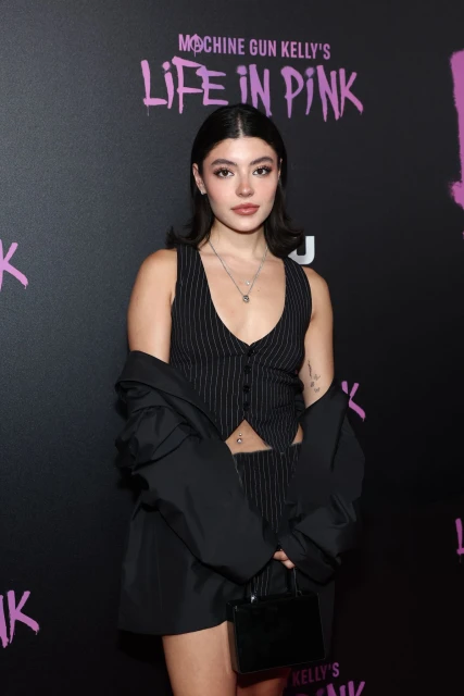 A fashionable young woman in a pinstripe vest and black jacket poses in front of a promotional backdrop. She holds a small purse.