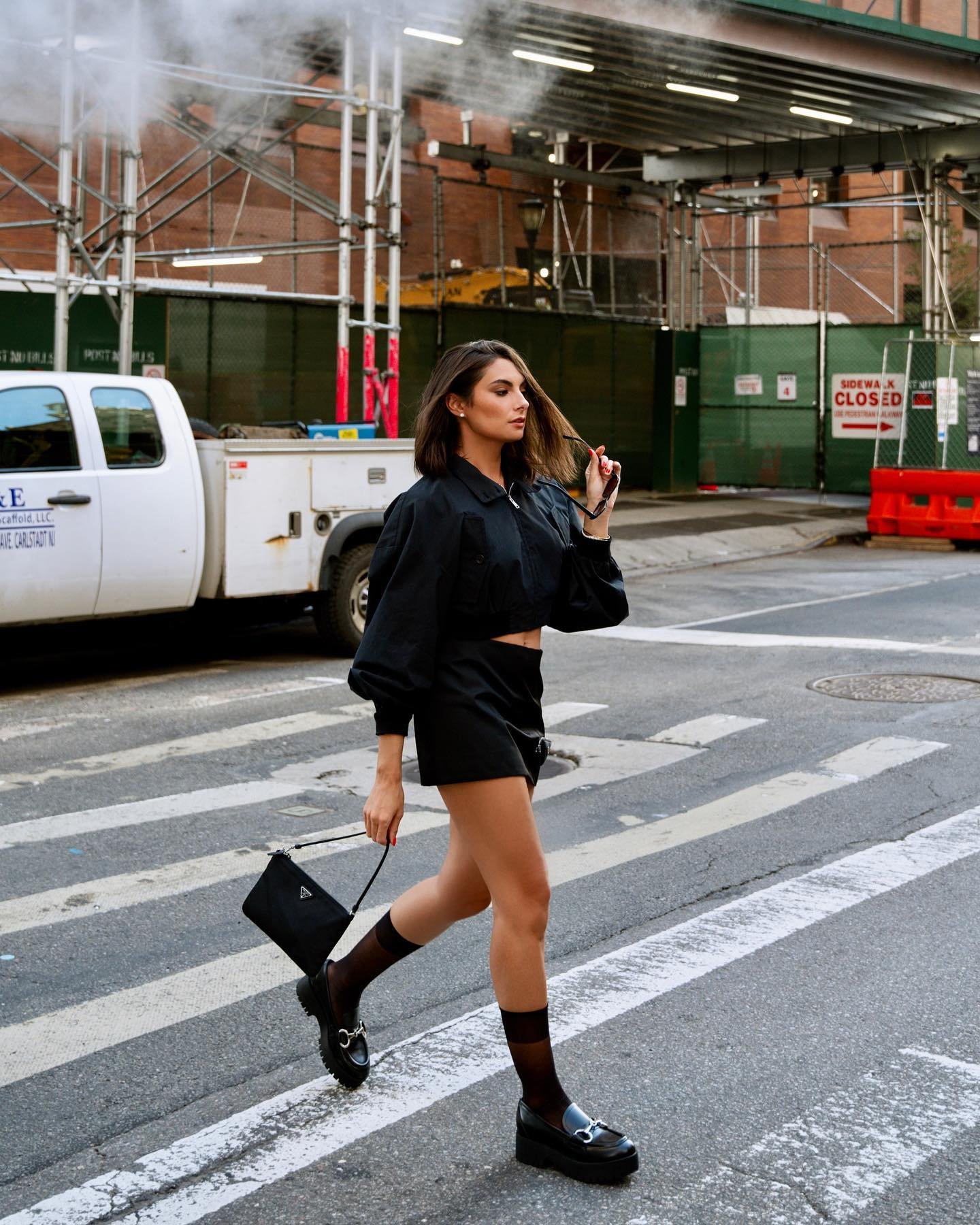 Woman in black outfit crosses urban street in daytime, holding Prada bag, wearing loafers and sheer socks, with a neutral expression.
