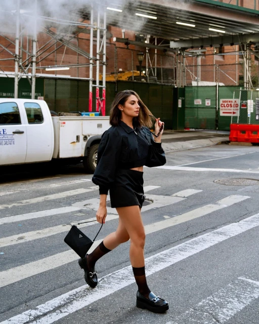 Woman in black outfit crosses urban street in daytime, holding Prada bag, wearing loafers and sheer socks, with a neutral expression.