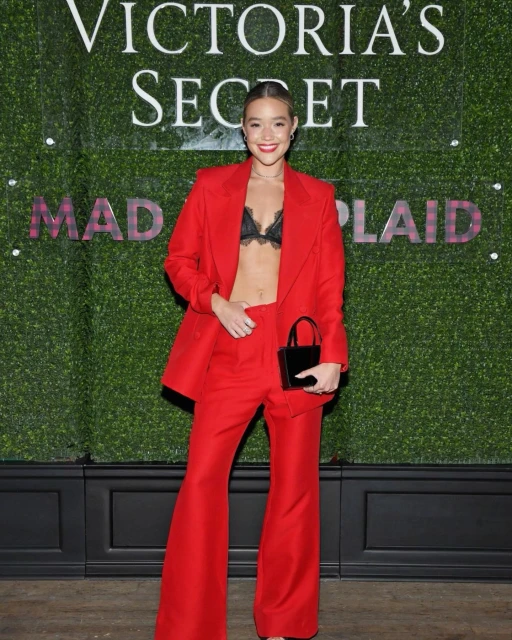Young woman in red pantsuit and black lace bra top poses in front of Victoria's Secret logo and greenery wall at event.