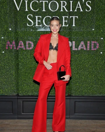 Young woman in red pantsuit and black lace bra top poses in front of Victoria's Secret logo and greenery wall at event.