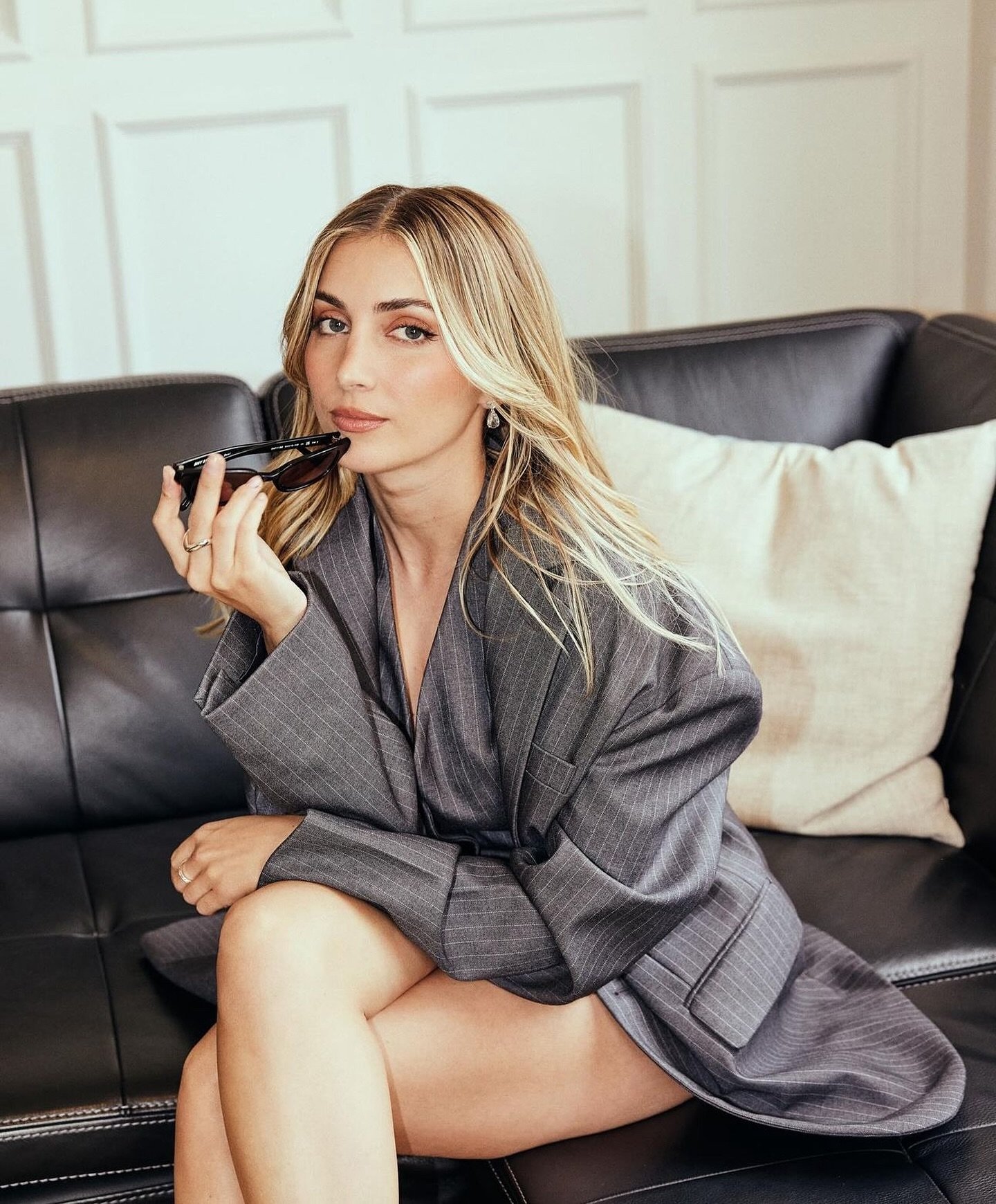 Blonde woman in oversized pinstripe suit jacket poses on a black leather sofa holding sunglasses with a white wall background.
