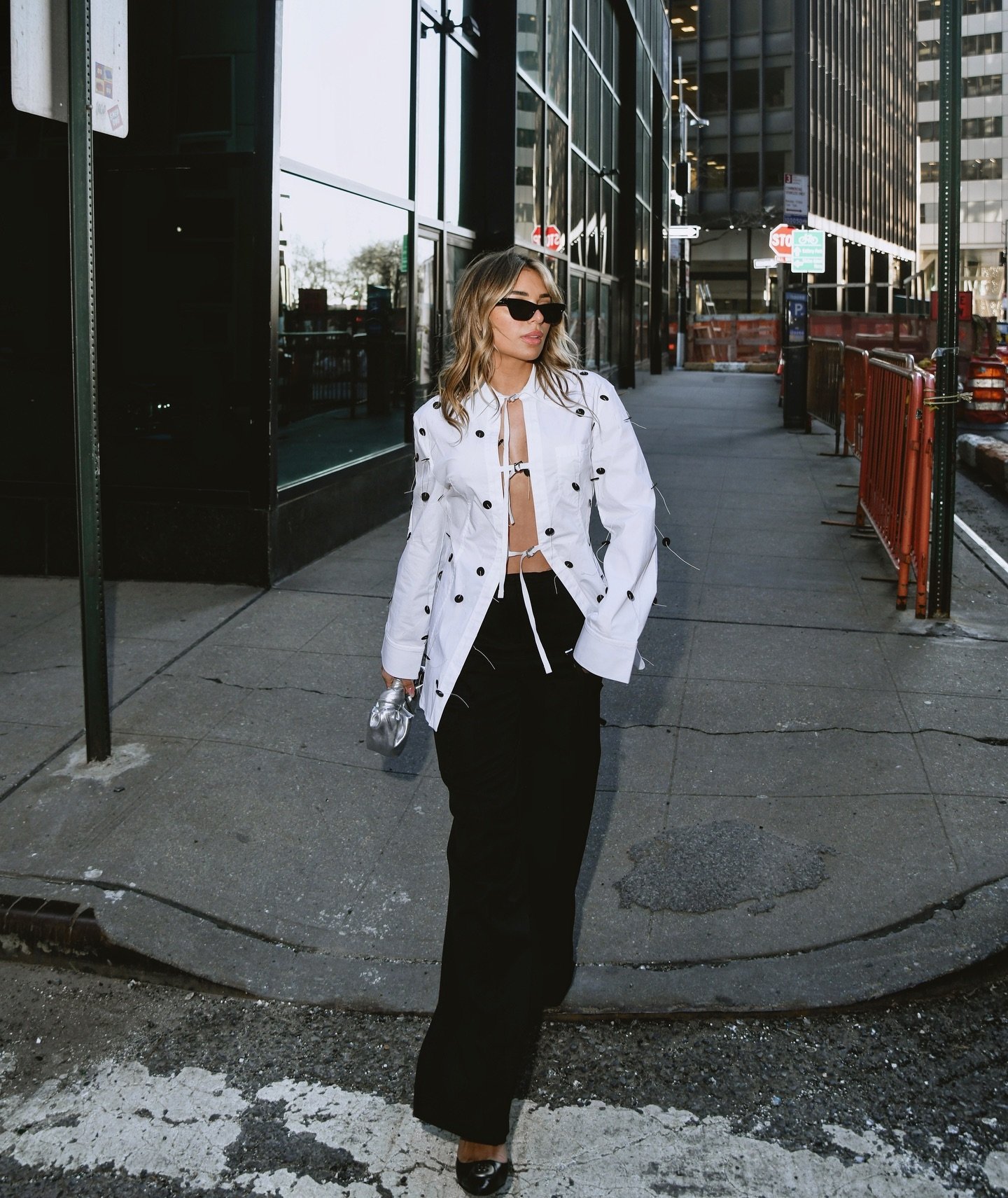 Adult female walks down urban sidewalk in white jacket with black dots and wide-leg black pants, holding water bottle on sunny day.