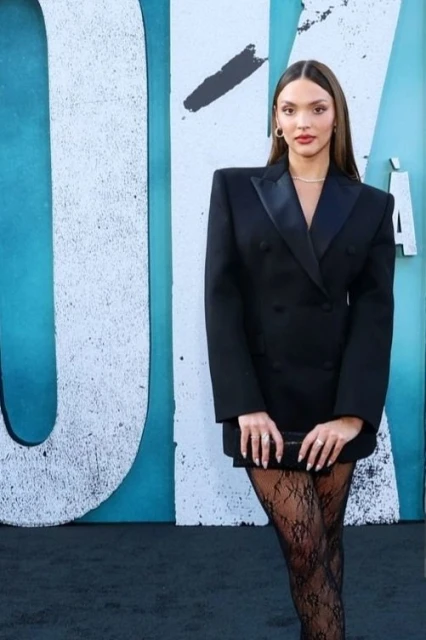 Elegant woman in a black double-breasted blazer dress and lace stockings poses in front of a graffiti-covered wall at an event.