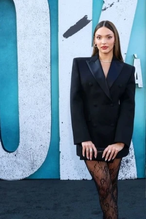 Elegant woman in a black double-breasted blazer dress and lace stockings poses in front of a graffiti-covered wall at an event.