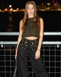 Woman wearing a black studded sleeveless top and matching trousers posing against a metal railing at night.