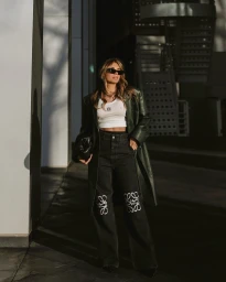 Woman wearing dark sunglasses, a green leather coat, white top, and patterned black jeans holding a black clutch in an urban setting