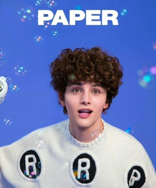 Young male model with curly hair wears a lettered sweater in a portrait-style close-up against a blue background with floating bubble effects.