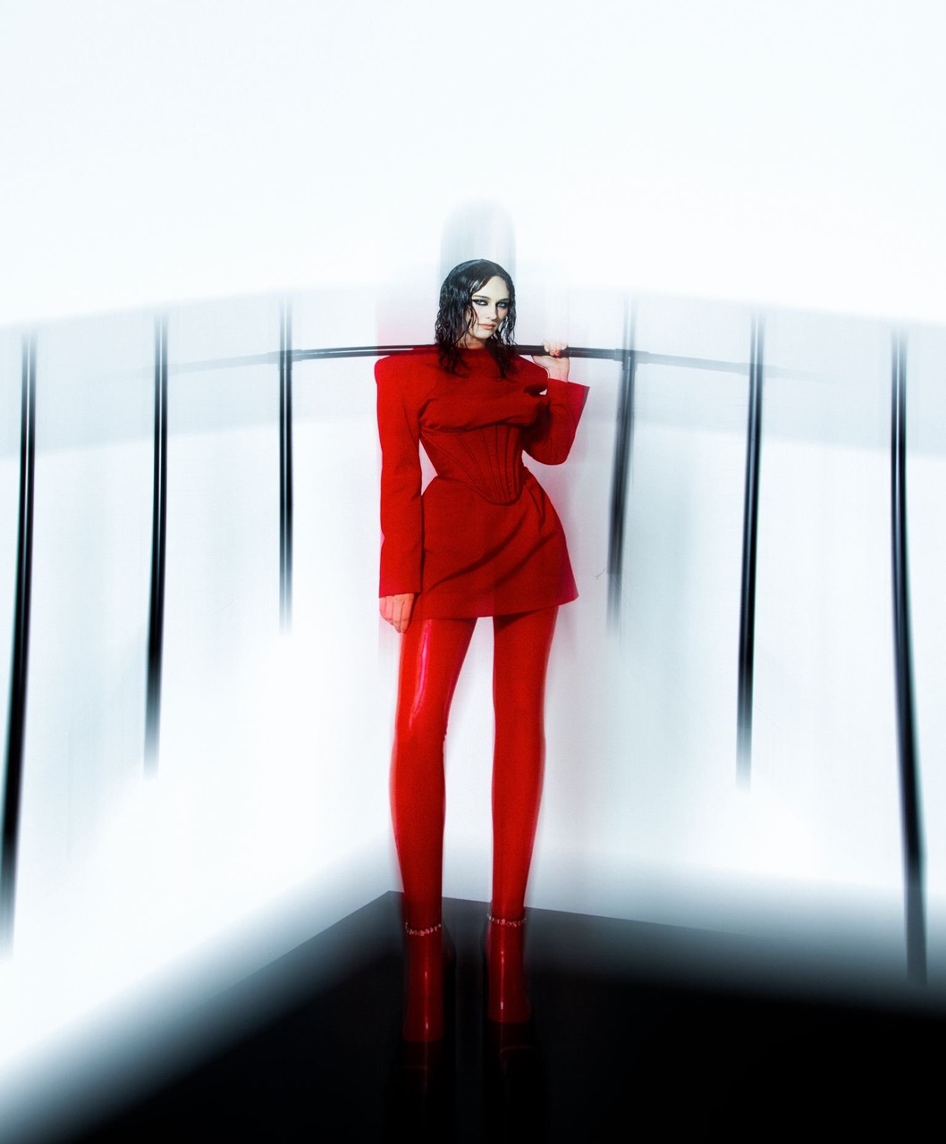 A woman in a red dress and leggings poses with a metal bar in a studio setting with white walls and a black floor.