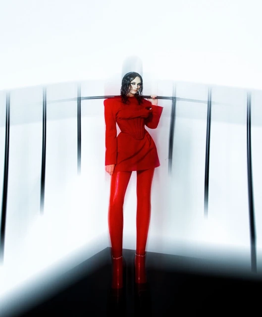 A woman in a red dress and leggings poses with a metal bar in a studio setting with white walls and a black floor.