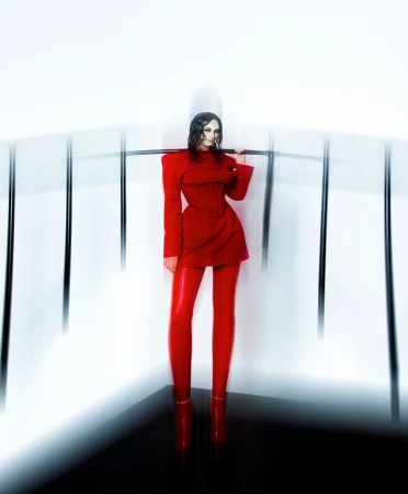 A woman in a red dress and leggings poses with a metal bar in a studio setting with white walls and a black floor.