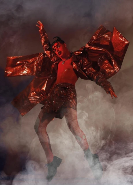 Person with dramatic red makeup and shiny red clothing leaps amid smokey background in a visually striking, fashion-forward studio shot.