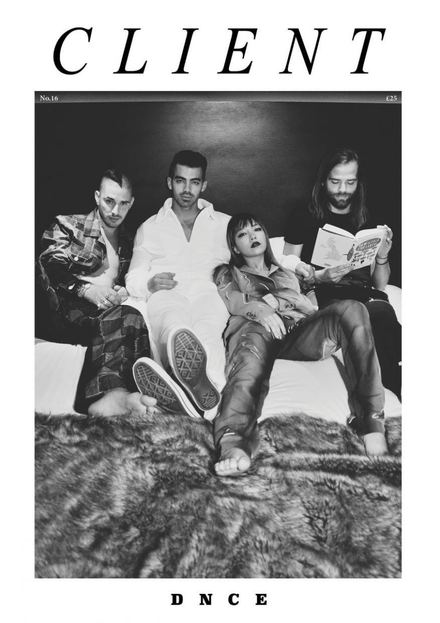 Black and white image features four young adult band members sitting on a fur-covered bed, one reading, in a magazine cover style.
