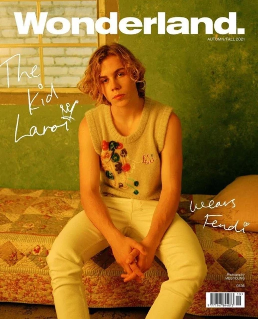 Magazine cover featuring The Kid Laroi. He's seated in a colorful knit sweater and cream pants against a muted green backdrop with a window.