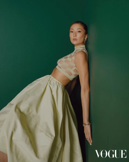 An Asian woman in a beige crop top and skirt leans against a dark green wall in a fashion editorial shot, gazing directly at the viewer.