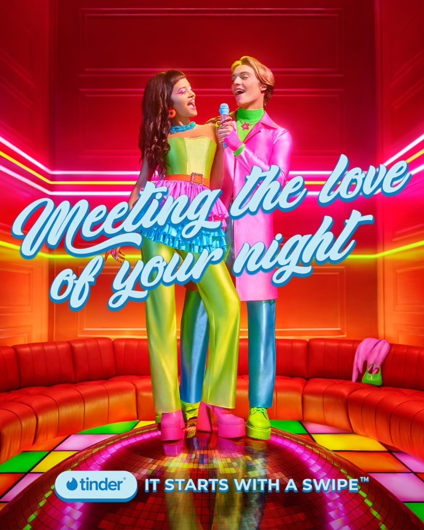 A woman and a man stand on a disco ball, singing into a microphone in a retro-themed room with neon lights and orange seating.