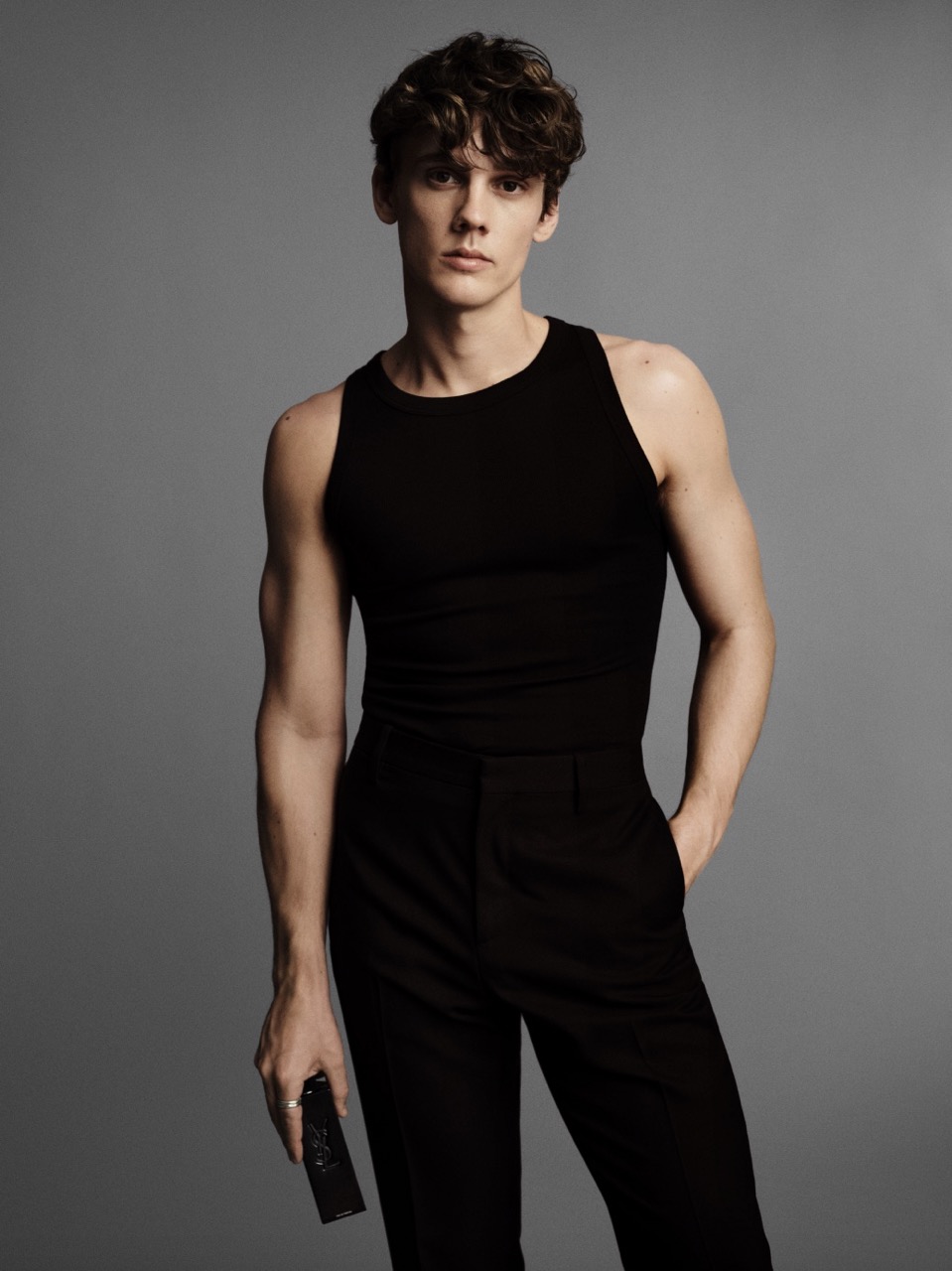 A young adult male in a black tank top and black pants poses in a studio, holding a black object. The background is neutral gray.