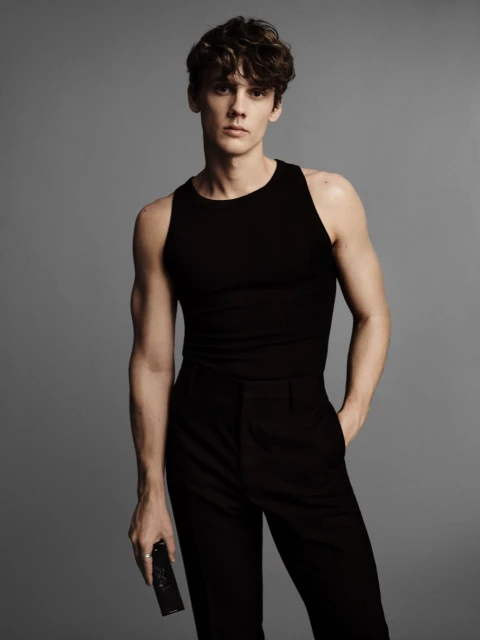 A young adult male in a black tank top and black pants poses in a studio, holding a black object. The background is neutral gray.