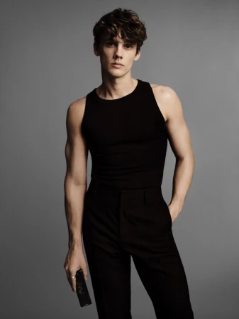 A young adult male in a black tank top and black pants poses in a studio, holding a black object. The background is neutral gray.