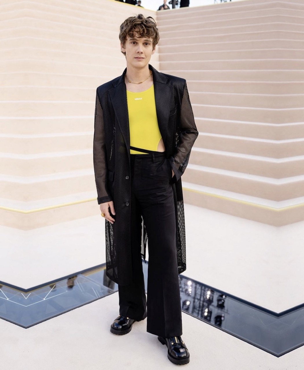 Young male model in a black mesh coat over a yellow top and black pants poses on stairs at a fashion event with neutral tones.