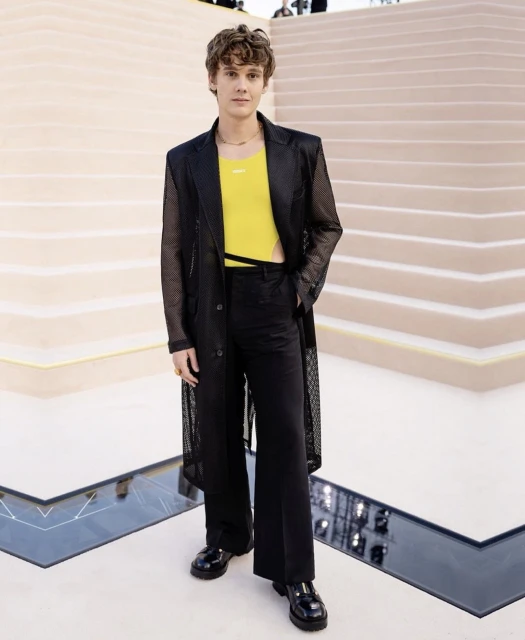 Young male model in a black mesh coat over a yellow top and black pants poses on stairs at a fashion event with neutral tones.