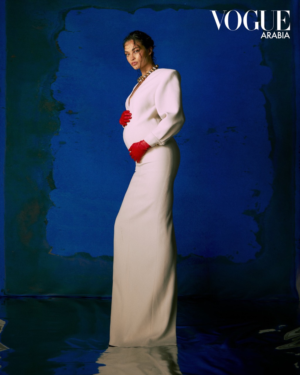 A pregnant woman wearing a full-length white gown and red gloves stands against a blue painted backdrop in a fashion editorial shot.