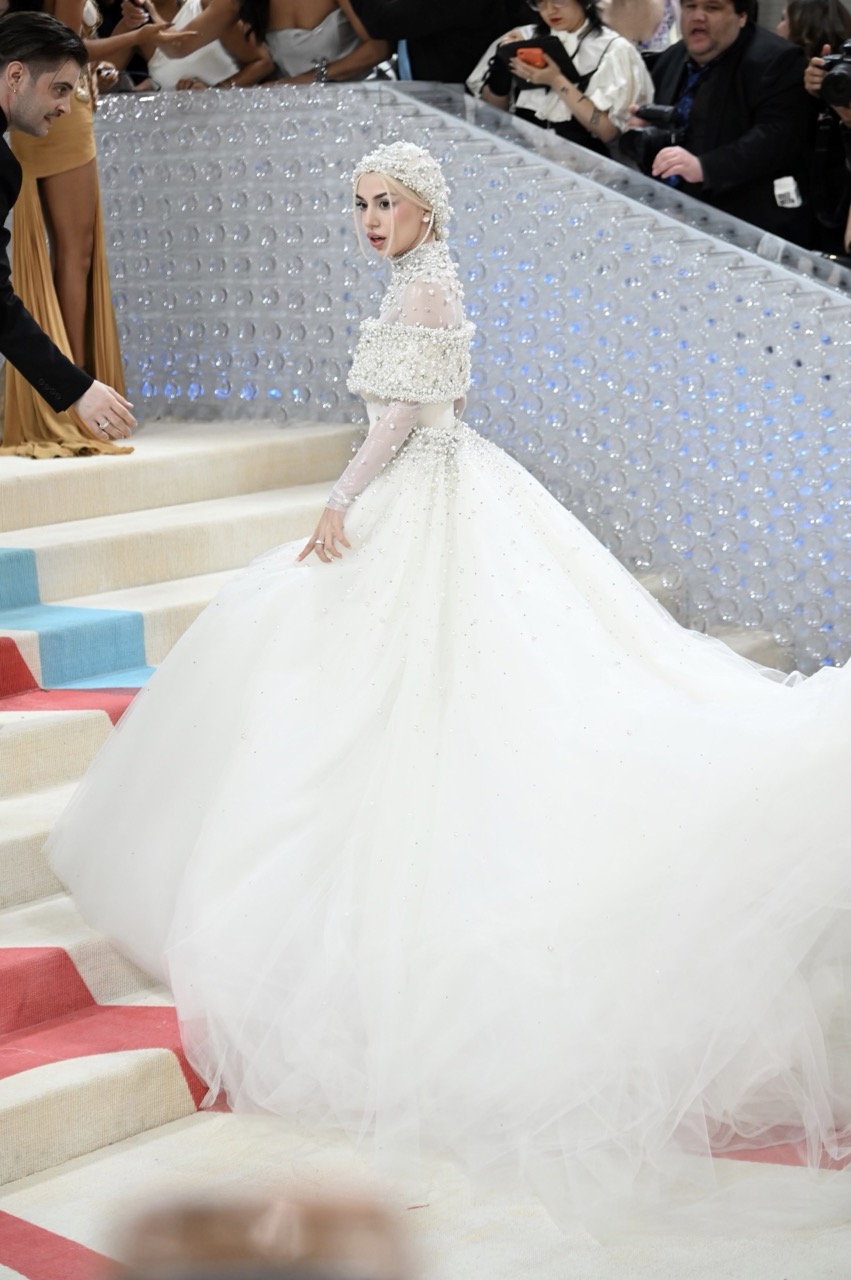 Woman in a white beaded wedding dress walks down a red and blue striped staircase at a fashion event with onlookers in the background.