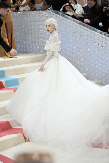 Woman in a white beaded wedding dress walks down a red and blue striped staircase at a fashion event with onlookers in the background.