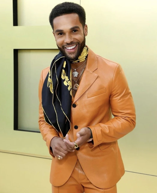 Smiling adult male in an orange leather suit with a Versace scarf and gold jewelry standing indoors against a neutral yellow background.