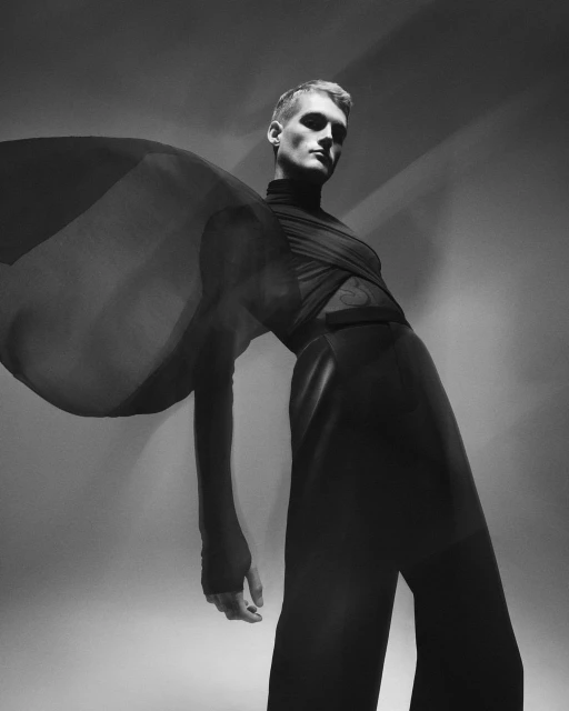 Monochromatic fashion shot of a male model in a draped black outfit, striking a pose in a studio setting, with a light gray background.