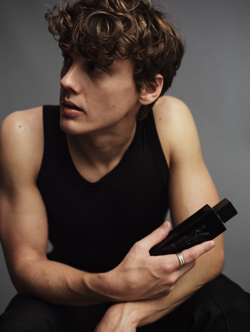 Young man with curly brown hair wearing a black tank top holding a dark perfume bottle against a grey background