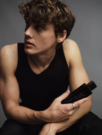 Young man with curly brown hair wearing a black tank top holding a dark perfume bottle against a grey background