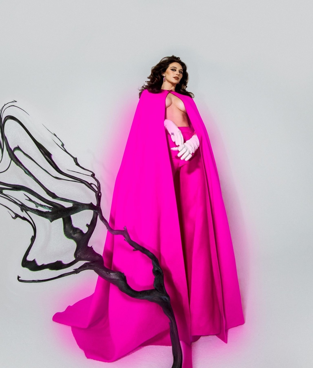 Model in vibrant hot pink cape and matching trousers holding white gloves against a plain white studio background.