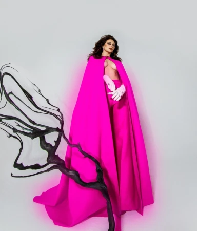 Model in vibrant hot pink cape and matching trousers holding white gloves against a plain white studio background.