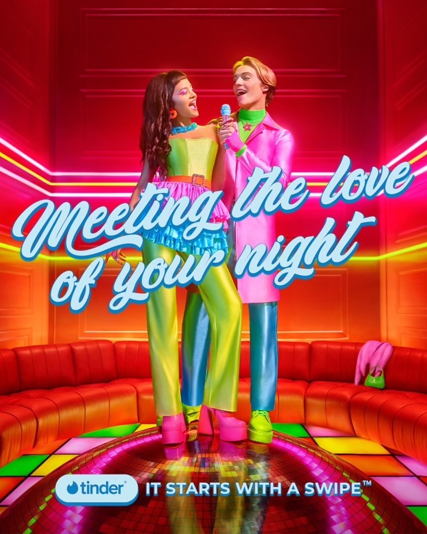 A man and a woman are singing together on a stage with a disco ball, dressed in bright retro 1970s inspired clothes and matching platform shoes.