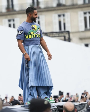 Man standing on a car roof wearing a blue short-sleeve sweater and a long pleated skirt by Danyul Brown.