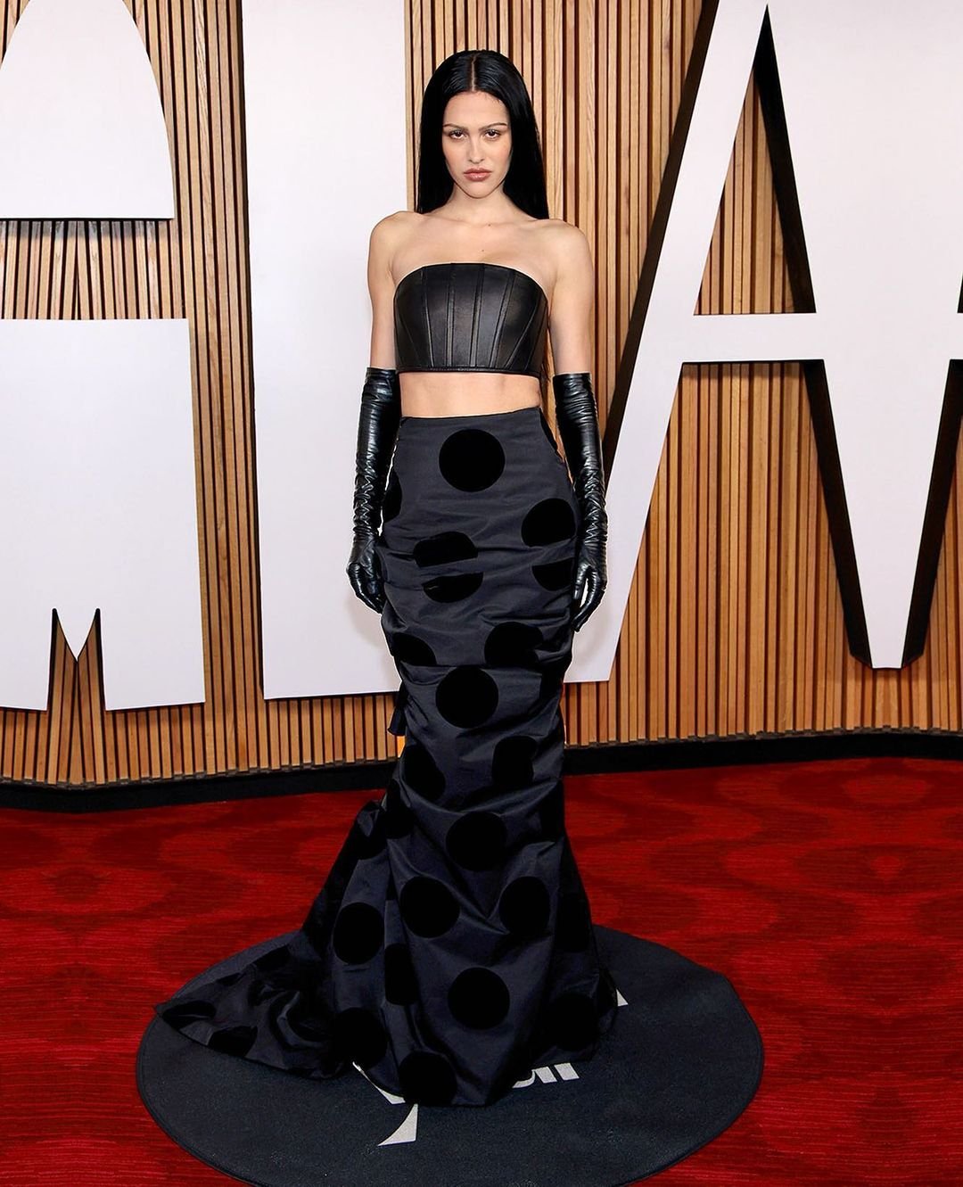 An adult woman poses in a strapless top, long gloves, and polka dot skirt on a red carpet in front of an interior wood background.