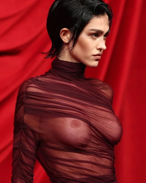 A woman with short black hair and a serious expression wears a sheer brown top against a solid red background in a fashion-style portrait.