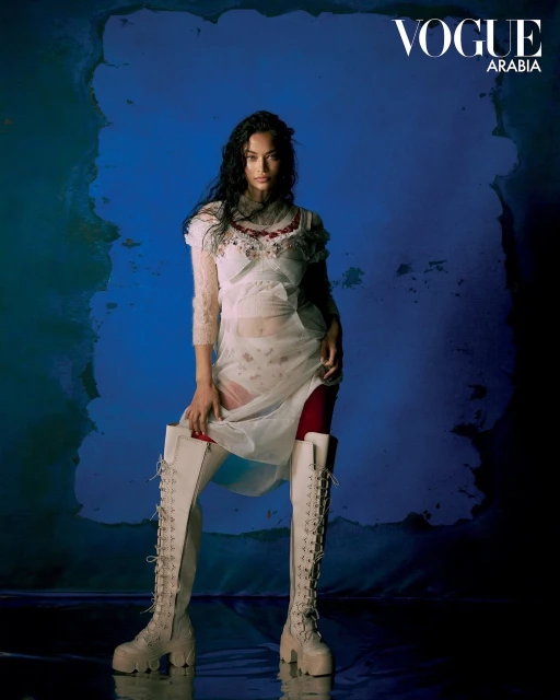 Fashion portrait of a woman posing in a white dress and tall lace-up boots against a blue textured backdrop with studio lighting.