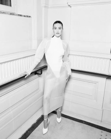 Monochrome portrait of a woman in a fitted white dress and heels standing against an ornate wall with paneling.