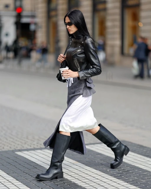 Brunette woman in sunglasses and a leather jacket walks across a crosswalk carrying a coffee cup in an urban environment.