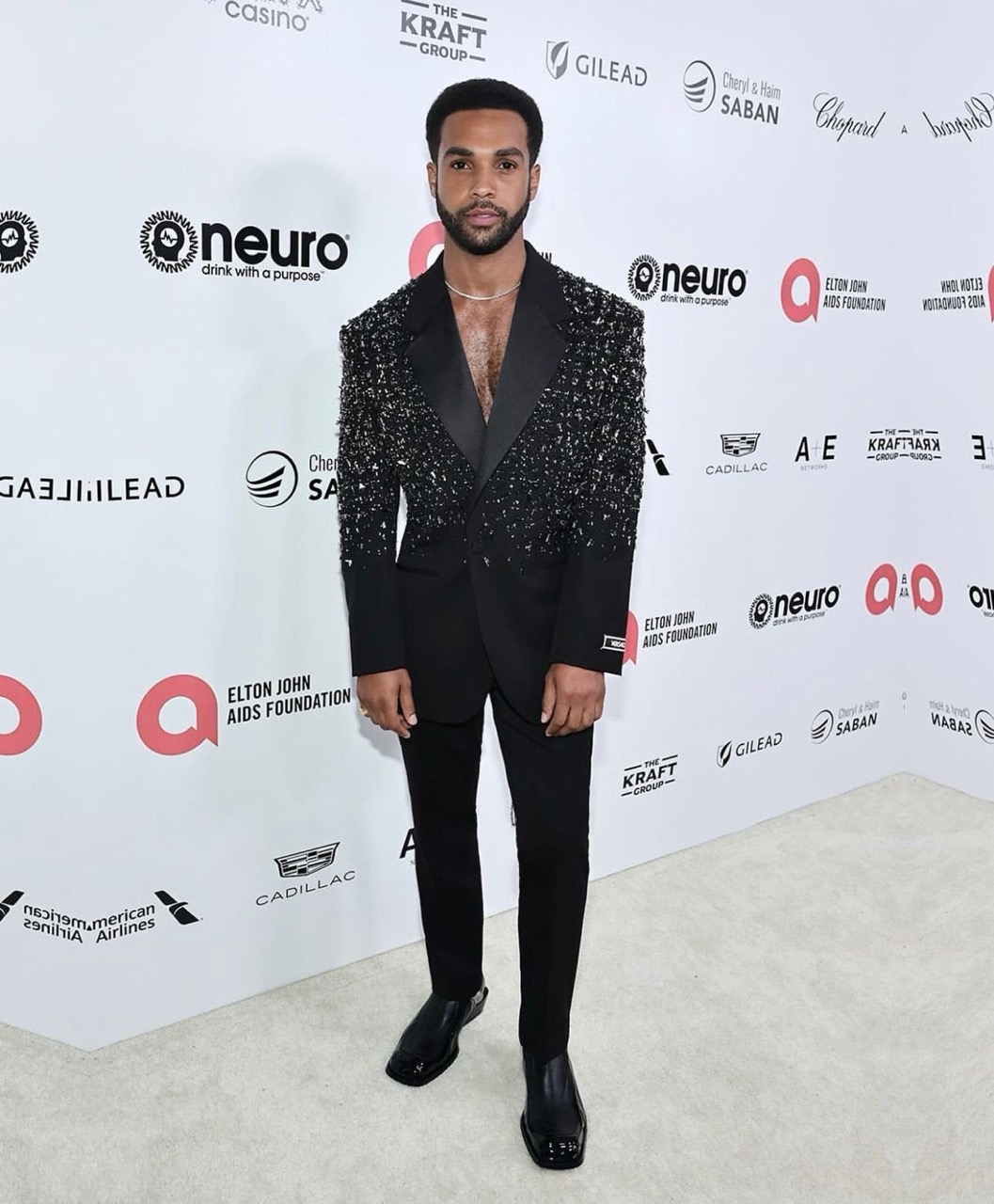 Full-length portrait of a Black man in a sparkling black suit at an Elton John AIDS Foundation event on a white patterned carpet.
