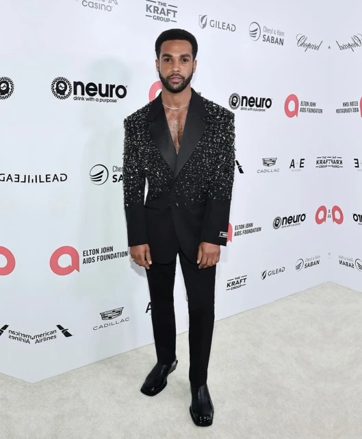 Full-length portrait of a Black man in a sparkling black suit at an Elton John AIDS Foundation event on a white patterned carpet.