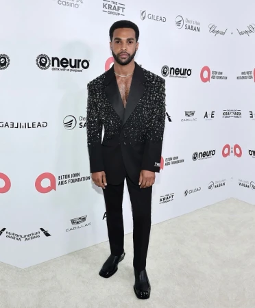 Full-length portrait of a Black man in a sparkling black suit at an Elton John AIDS Foundation event on a white patterned carpet.