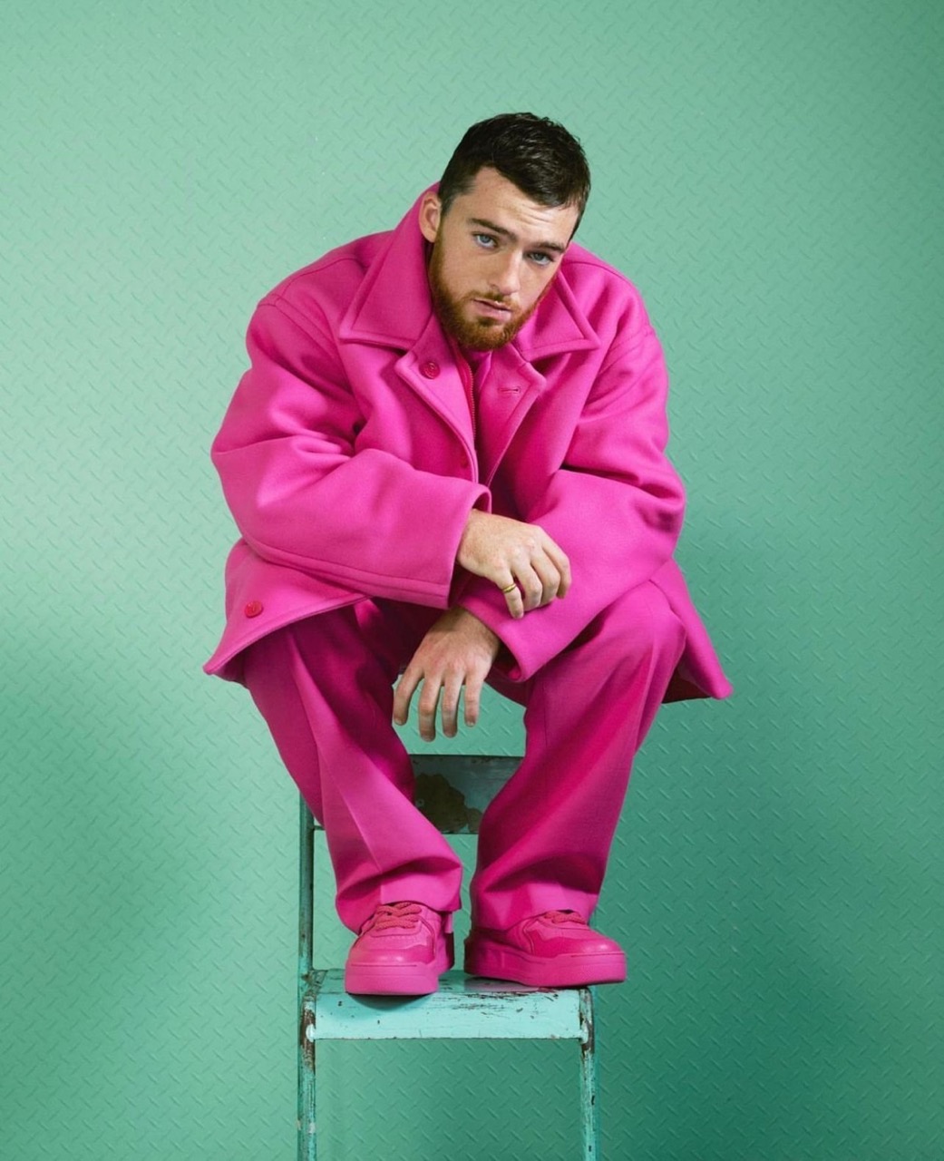 Portrait of a man in a bright pink suit sitting on a blue metal step stool against a light green textured background, eye-level shot.